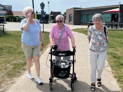 Drei Seniorinnen gehen an einem sonnigen Tag , wobei eine Person einen Rollator benutzt, spazieren.